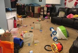 Toy room with toys all over the beige carpeted floor and  black couches