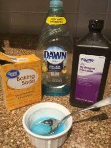 yellow box of baking soda, bottle of blue Dawn dish detergent, brown bottle of hydrogen peroxide, small white dish with blue mixture inside and a spoon.