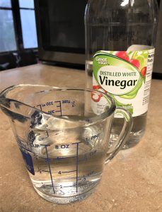 Glass measuring cup with vinegar in it with a bottle of vinegar behind on a counter top.